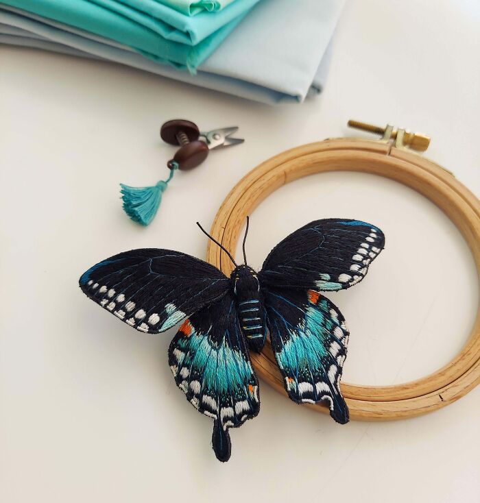 Creative embroidery of a butterfly with teal and black wings next to sewing tools and fabric.