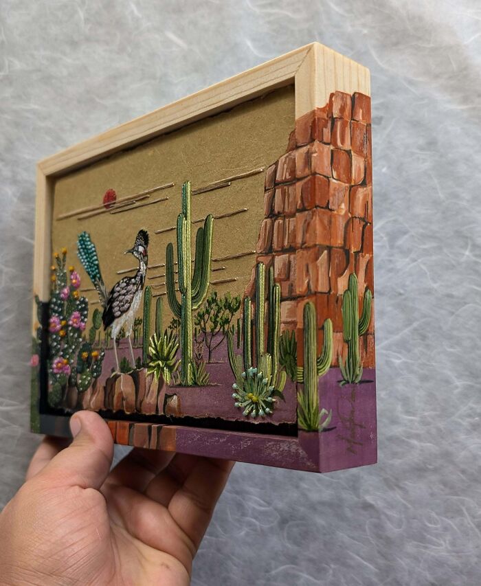 Creative embroidery of a desert scene with cacti and rocks, displayed in a wooden frame.