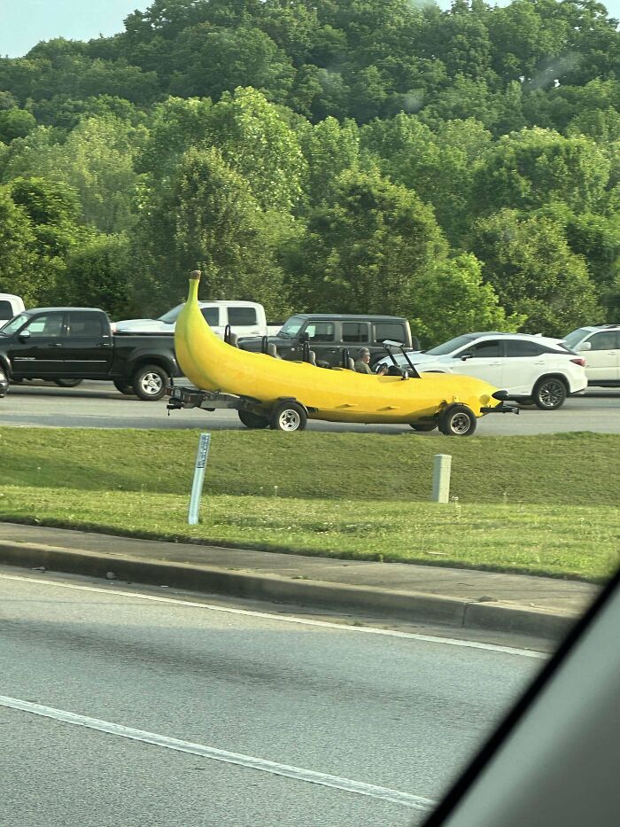 Yellow banana-shaped car on the road, surrounded by regular vehicles, with green trees in the background.