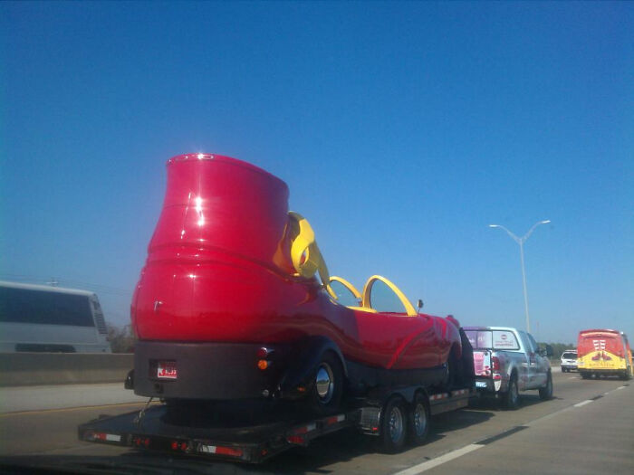 Giant shoe-shaped car on a trailer, spotted on the road under a clear blue sky.