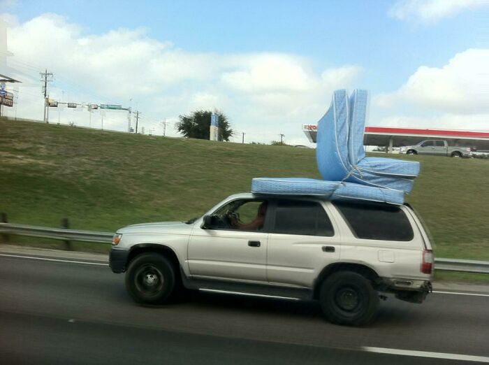 SUV on the road carrying a large stack of mattresses tied to the roof, highlighting interesting things seen while driving.