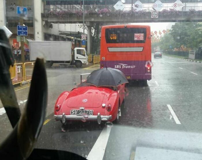 Red car with an umbrella on a rainy road, following a bus. Interesting things on the road captured in the city.