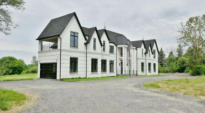 Large white mansion with black roof, showcasing that money can't buy taste, surrounded by a gravel driveway and trees.