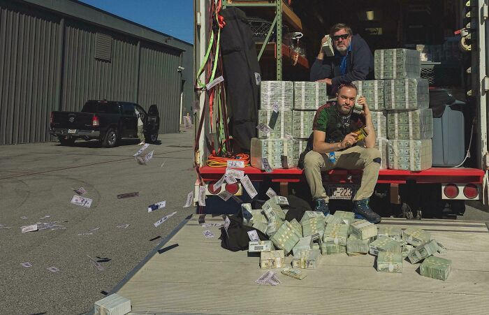 Two people sitting on a truck loaded with stacks of cash, with paper money scattered around, embodying good-funny-bosses vibe.