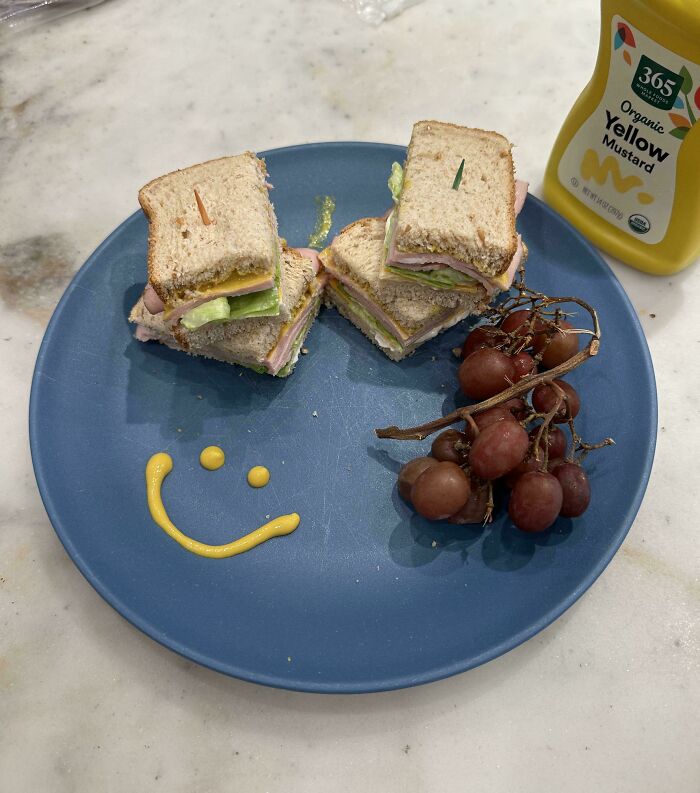 Plate with sandwich quarters, grapes, and a mustard smiley face; a fun parenting genius trick for kids' meals.