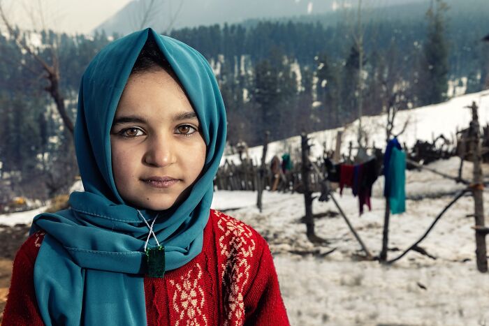 Young girl in a red sweater and blue headscarf standing in a snowy landscape, photographed by Hamed AlGhanboosi.