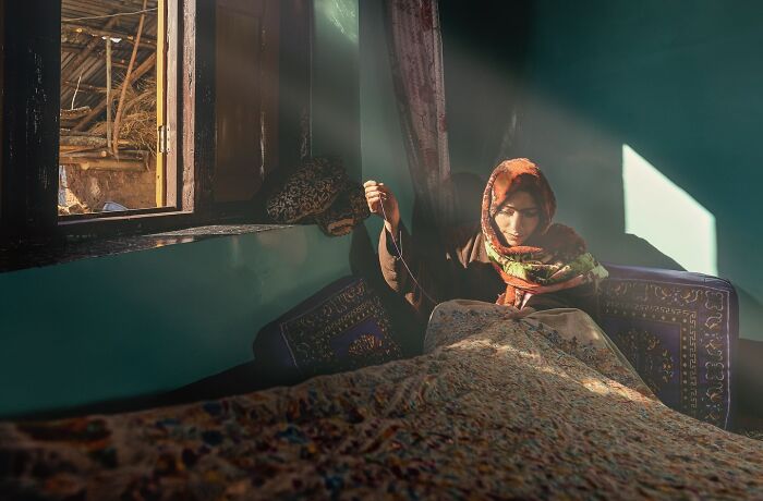 A woman sitting near a window, bathed in sunlight, weaving a carpet. Captivating photograph by Hamed AlGhanboosi.