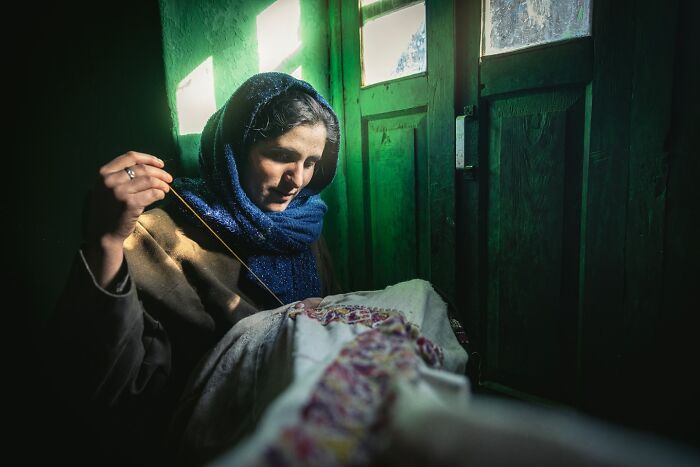 Woman sewing by a window in a warmly lit room, captured by Hamed AlGhanboosi.