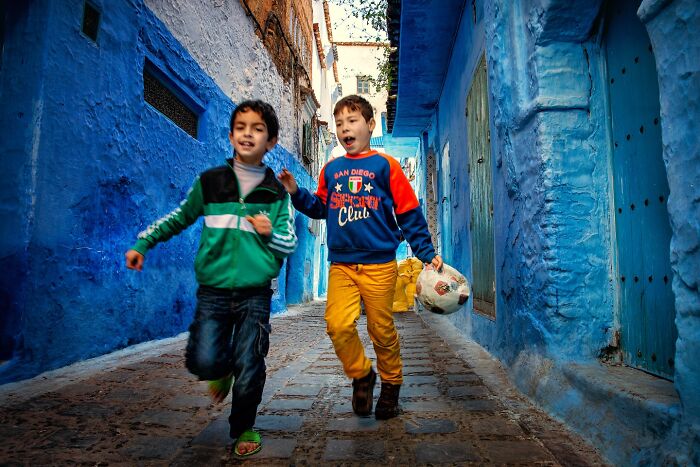 Two boys joyfully running through a vibrant blue alley, holding a soccer ball, captivated in Hamed AlGhanboosi's photograph.