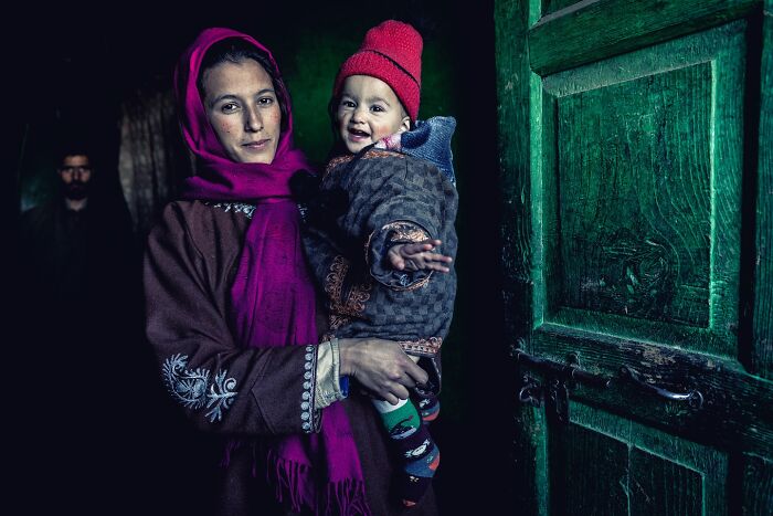 A woman in traditional attire holds a smiling child in a red hat, photographed by Hamed AlGhanboosi.