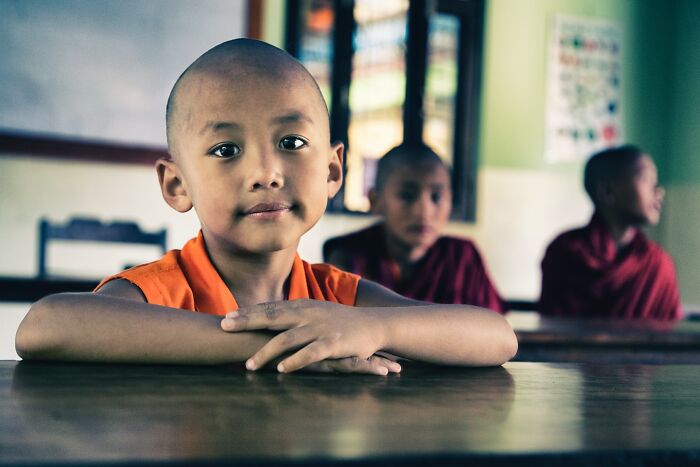 Young monk in classroom setting, photographed by Hamed AlGhanboosi, looking thoughtfully at the camera.