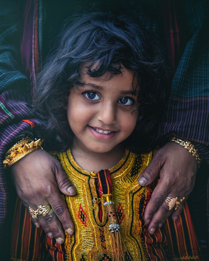 A captivating photograph by Hamed AlGhanboosi of a smiling child in colorful traditional attire, embraced by adorned hands.