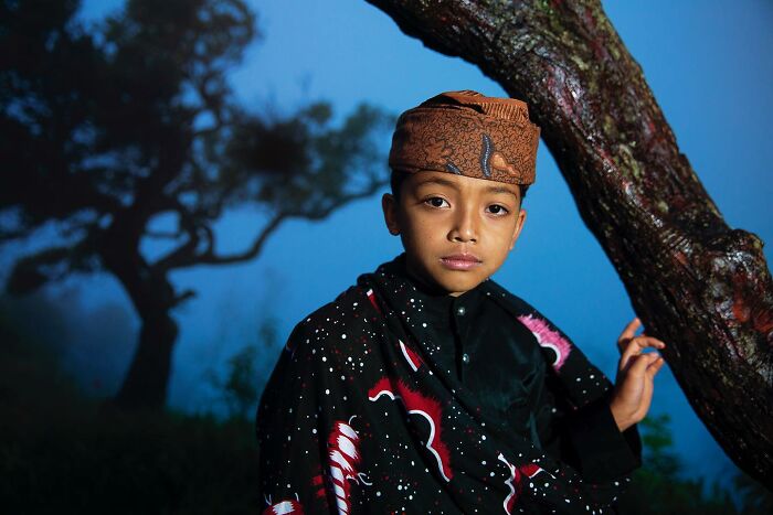 Young boy in traditional attire near a tree, vibrant nature backdrop; captivating photograph by Hamed AlGhanboosi.