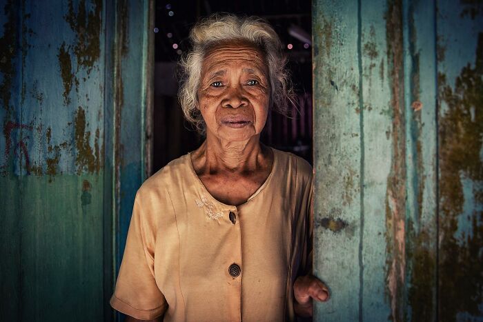 Elderly woman standing in a rustic doorway, captured in a captivating photograph by Hamed AlGhanboosi.