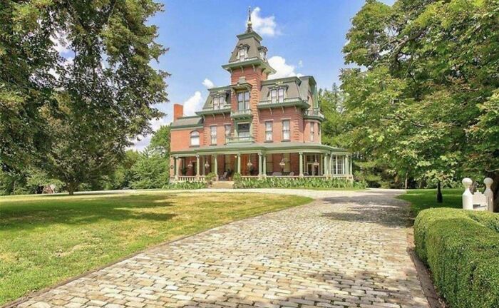 Victorian-style mansion with a long cobblestone driveway, surrounded by lush greenery, showcasing lack of taste despite wealth.