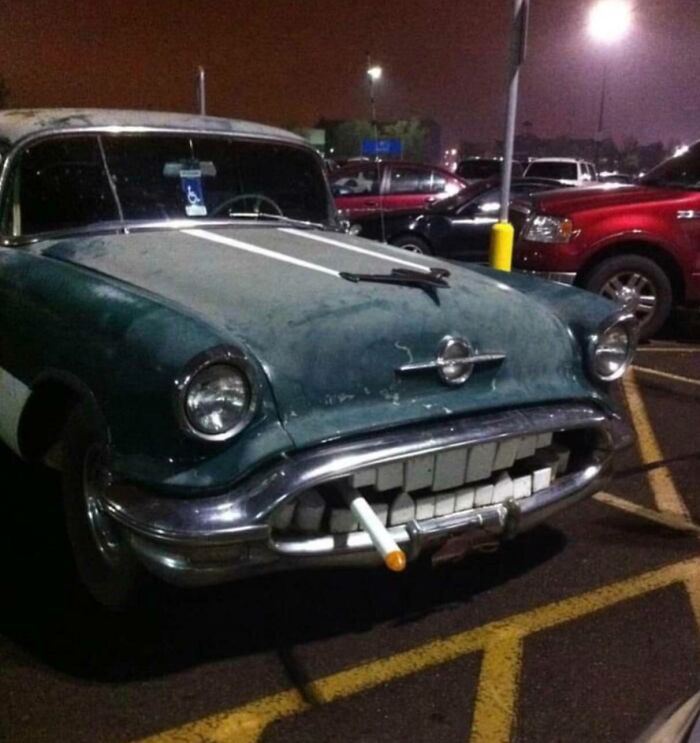 Classic car with a grill resembling teeth and a cigarette, highlighting a humorous car-fails scene in a parking lot at night.