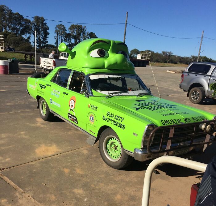 Green car fail with a large cartoon character face on the roof in a parking lot.