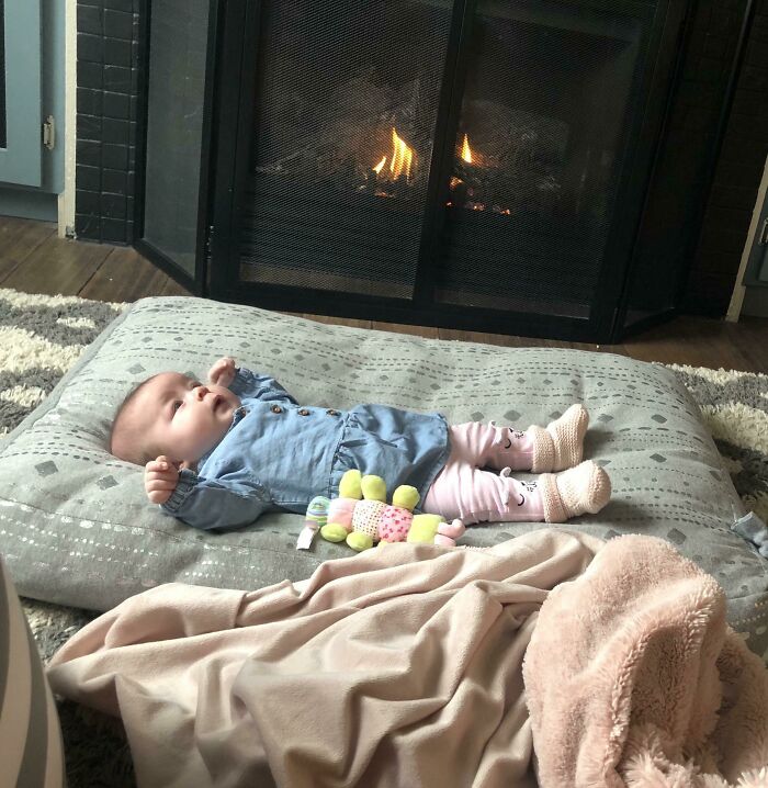 Baby lying on a cushion by a fireplace, showcasing cozy Parenting-Genius-Tricks for relaxation and warmth.