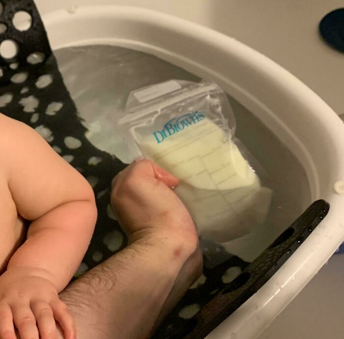 Baby in a bath with a parent's hand holding a milk bag, showcasing creative parenting tricks.