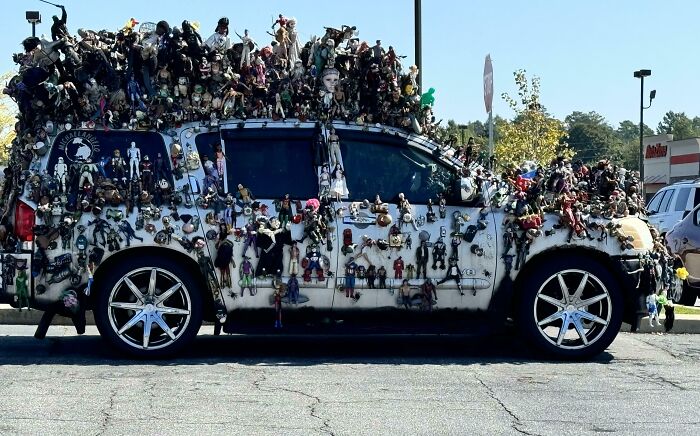 A car covered in numerous toy dolls, parked in a lot, showcasing an unusual decoration style.