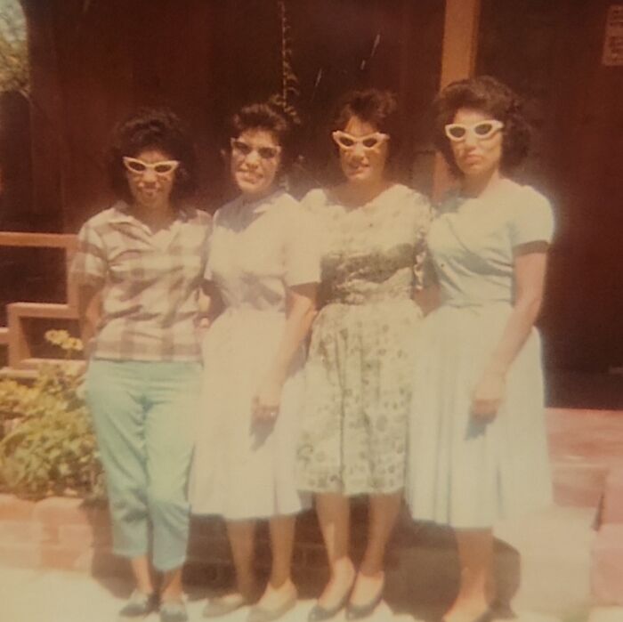 Four women in 60s fashion with retro sunglasses, standing together in vintage dresses and hairstyles.