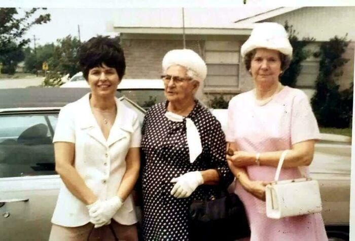 Three women in 60s fashion, wearing vintage dresses and hats, standing in front of a car.