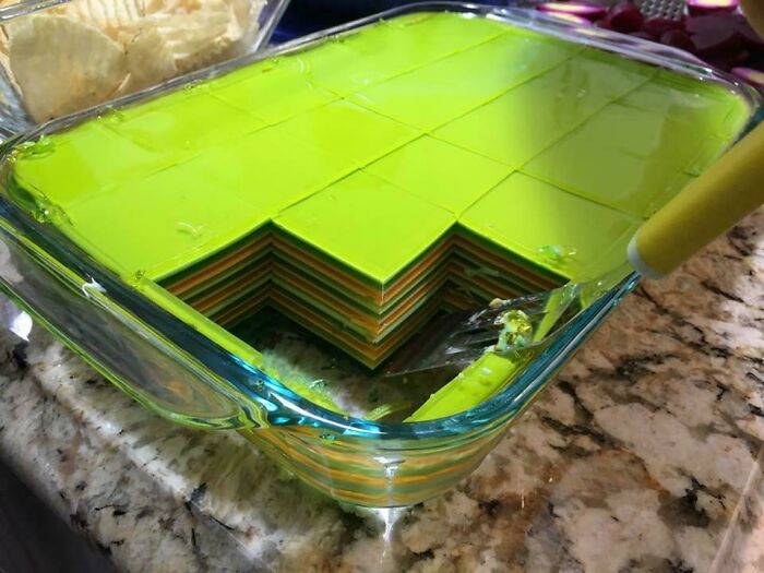 Colorful layered gelatin dessert in a glass dish with a piece cut out on a granite countertop. Fascinating pics.