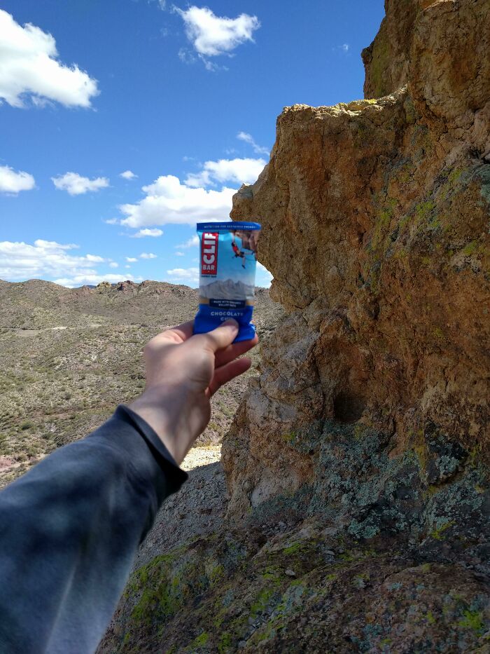 A hand holding a Clif Bar with a rocky desert landscape in the background under a blue sky with clouds.