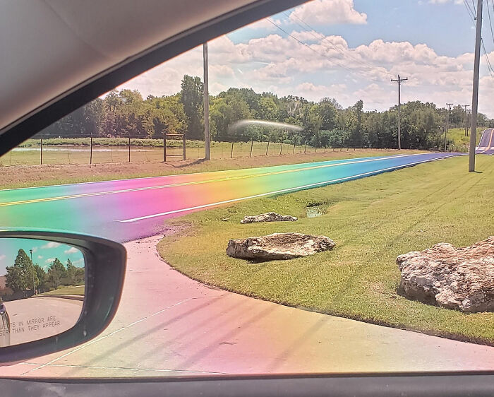 View from inside a car showing a fascinating rainbow-colored road under a clear blue sky with grassy surroundings.