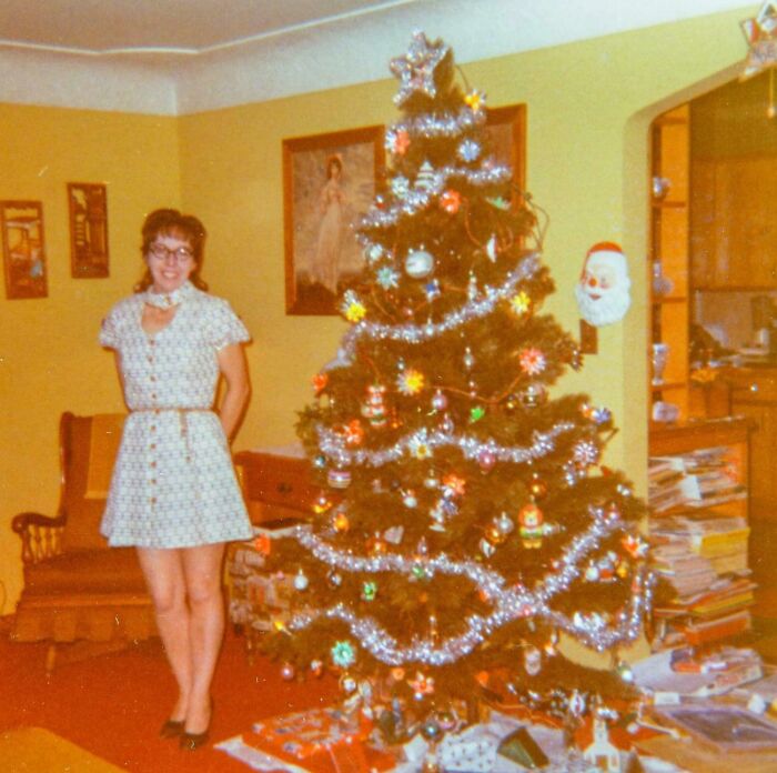 Woman wearing a 60s fashion dress standing next to a decorated Christmas tree in a vintage living room.