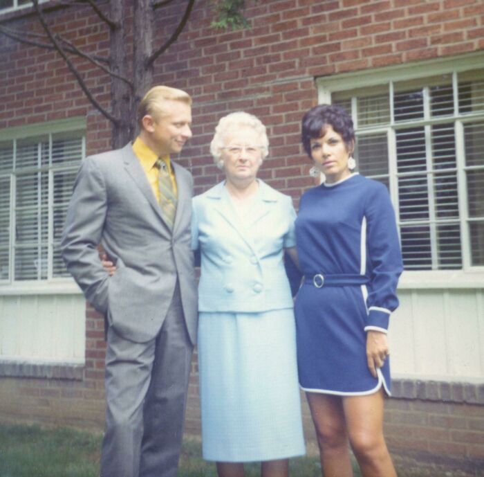 A woman in a blue 60s fashion dress, with two companions in front of a brick building.