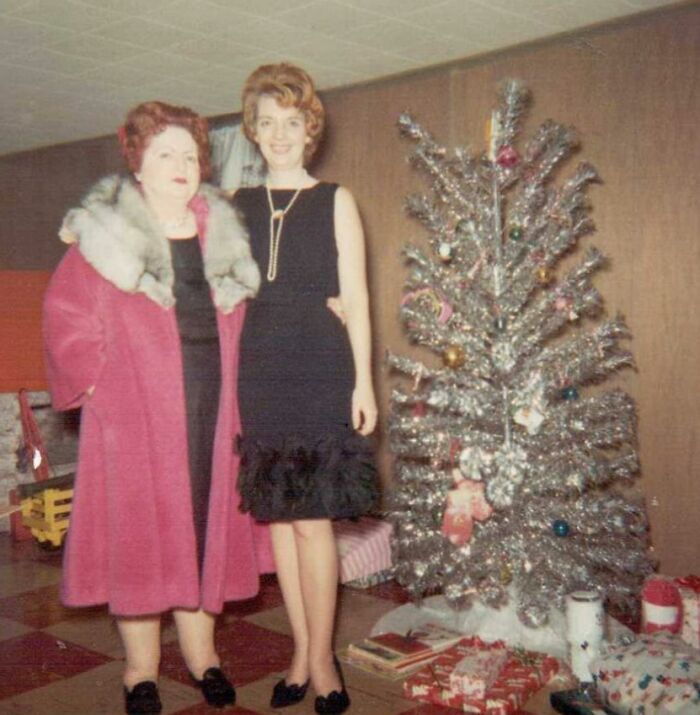 Two women in 60s fashion beside a vintage silver Christmas tree, one in a pink coat and the other in a black dress.