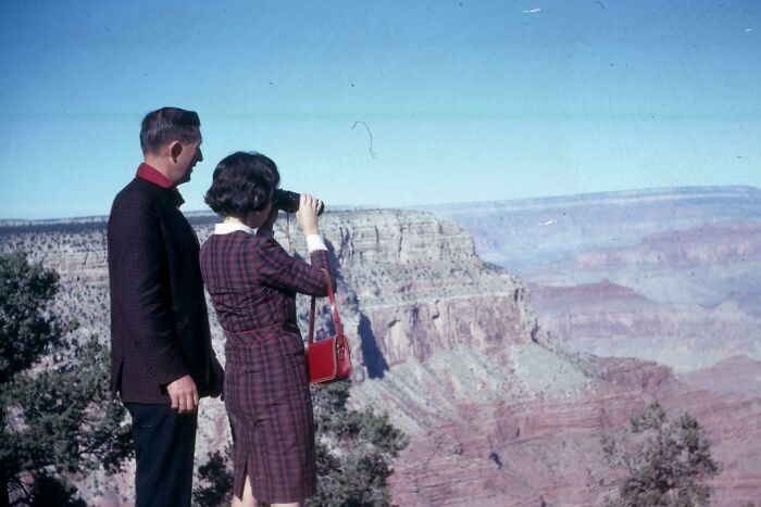 Couple in 60s fashion looks at Grand Canyon, woman with camera, wearing plaid dress and red handbag.