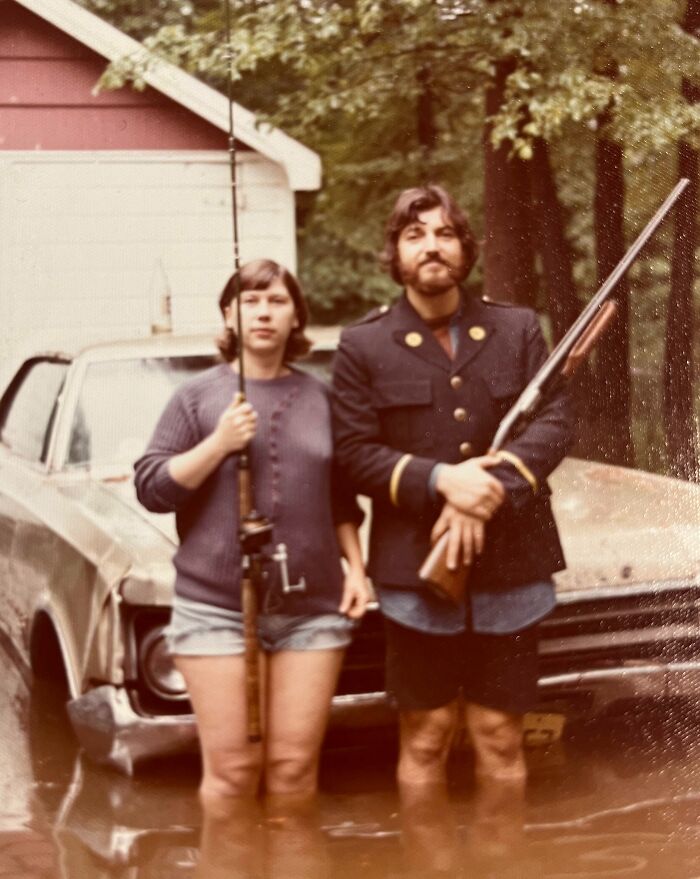 Two people in 60s fashion standing in front of a car, one holding a fishing rod, the other holding a rifle.