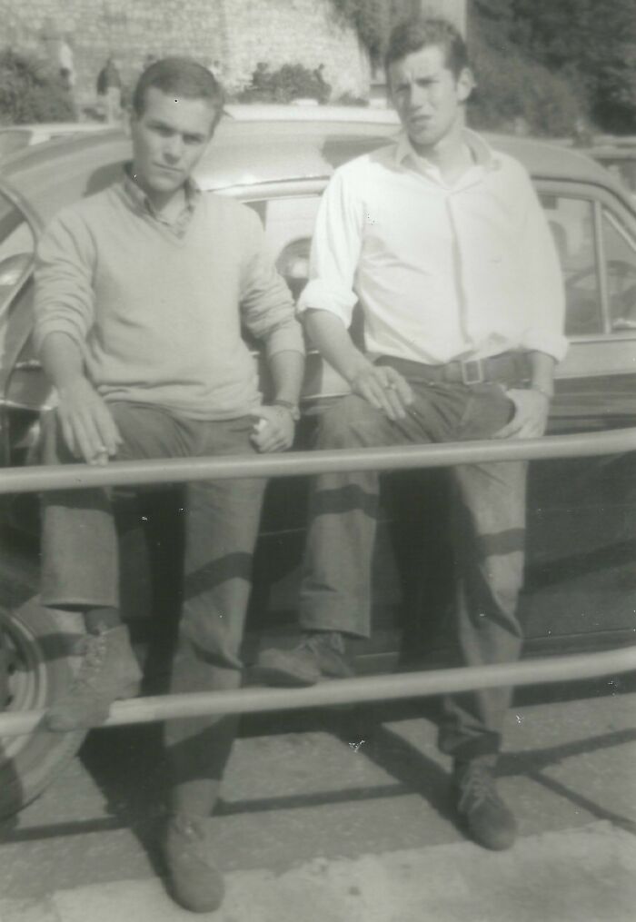 Two men in 60s fashion, standing by a vintage car, both wearing casual shirts and pants.