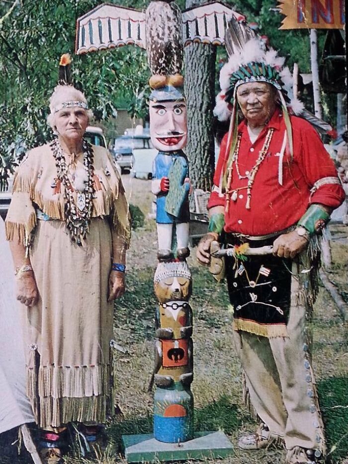 Two people in traditional attire from the 60s, standing next to a colorful totem pole.