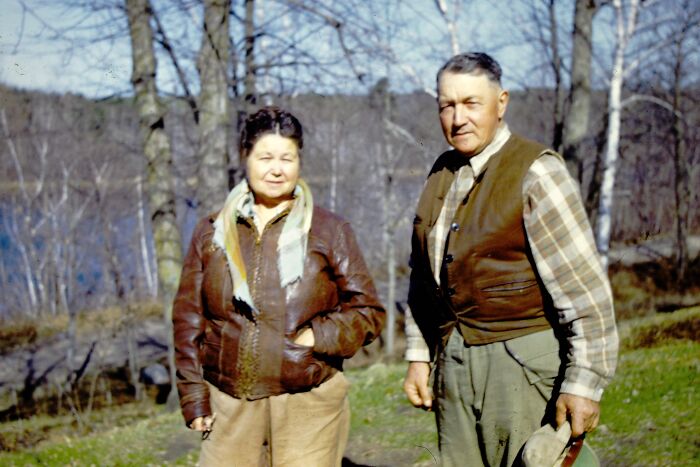 Elderly couple in 60s fashion looks with leather jackets and plaid shirts, standing outdoors in a forest setting.