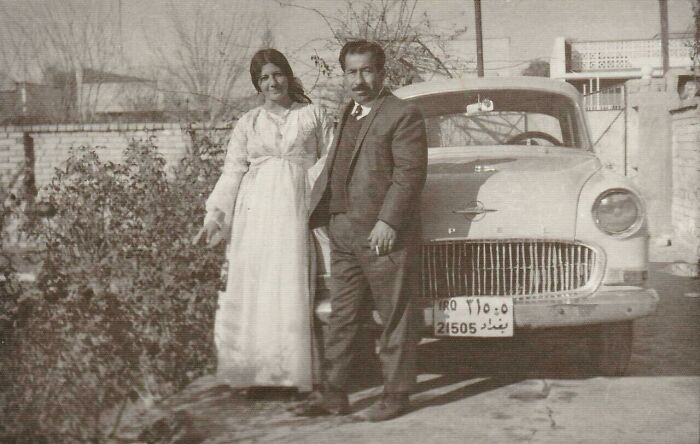 A couple in 60s fashion looks, with the woman in a long dress and the man in a suit, standing by a vintage car.