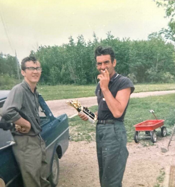 Two men in a 60s fashion look, standing by a car with a trophy and a red wagon nearby, outdoors.