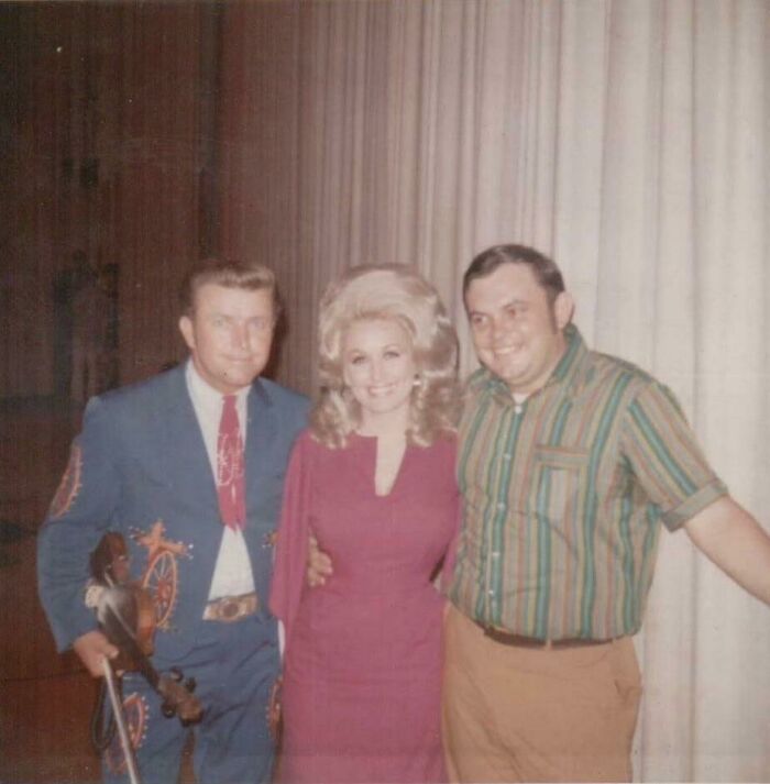 Three people in 60s fashion; a man in a blue suit, a woman with voluminous hair, and a man in a striped shirt, smiling together.