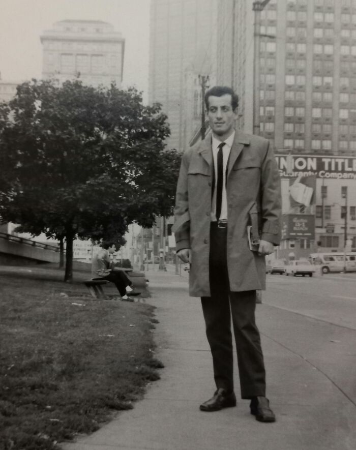 Man wearing a trench coat and tie on an urban sidewalk, exemplifying 60s fashion.