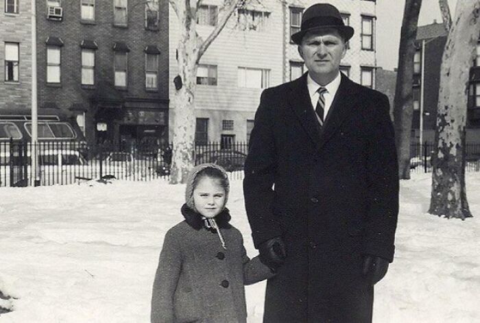 A man in a coat and hat with a young girl in a winter coat, showcasing 60s fashion, standing in a snowy urban setting.