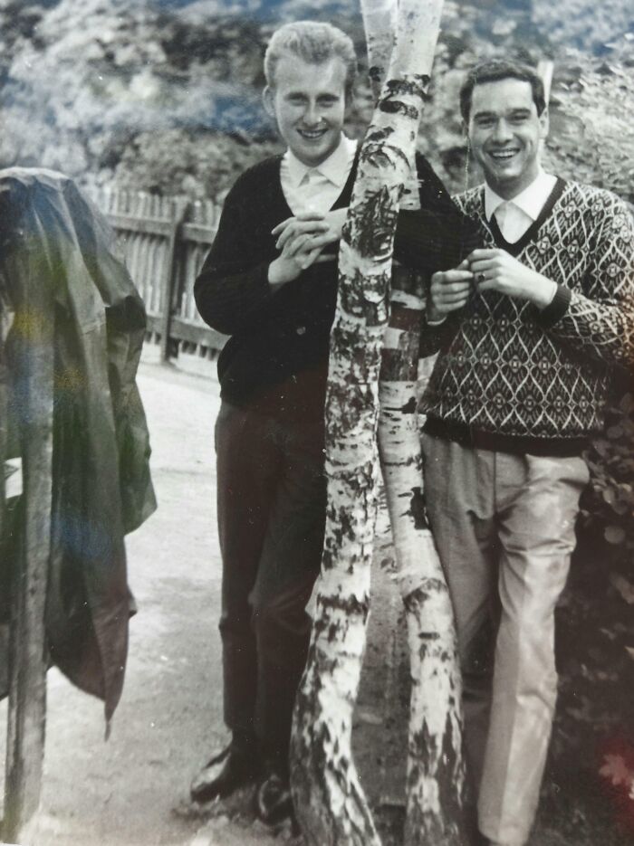 Two men in 60s fashion, one in a sweater, smiling by trees outdoors.