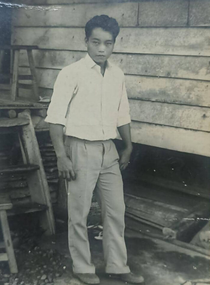 Man in 60s fashion look, wearing a white shirt and tailored trousers, posing outdoors by a wooden structure.