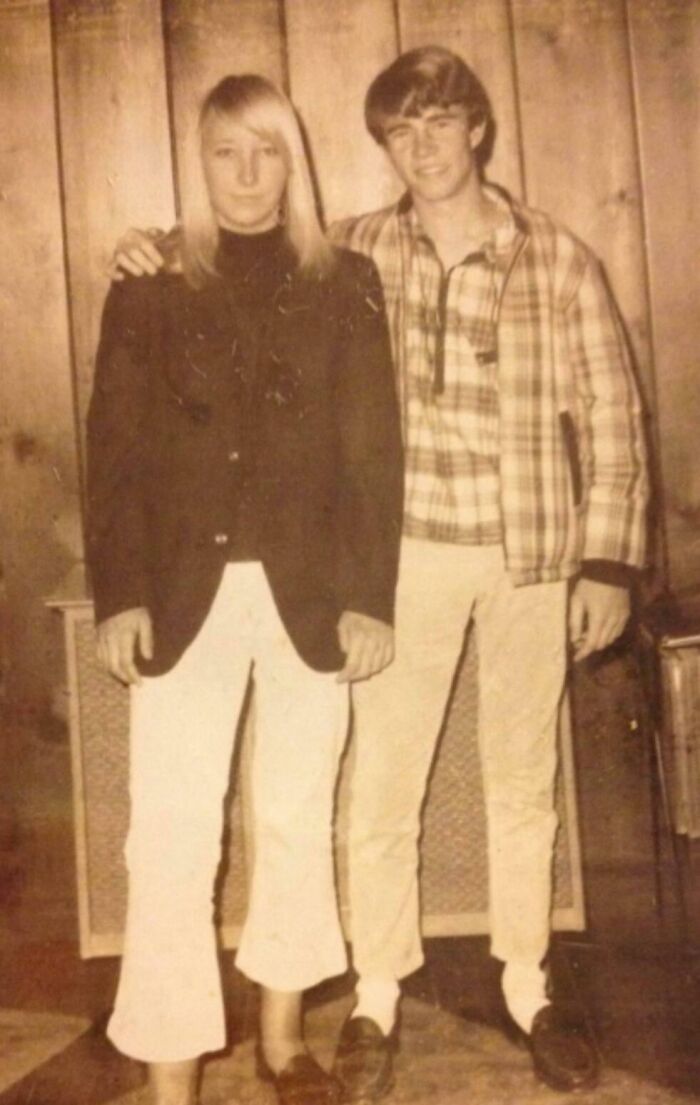 Two people in vintage 60s fashion, with flared pants and plaid jacket, posing together in a retro wood-paneled room.