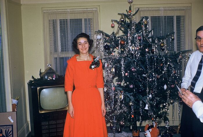 Woman in a 60s fashion look, wearing a red dress by a decorated Christmas tree, with a vintage TV in the background.