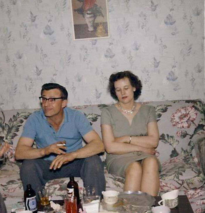 A couple sitting on a floral sofa, wearing 60s fashion, with a patterned wallpaper backdrop.