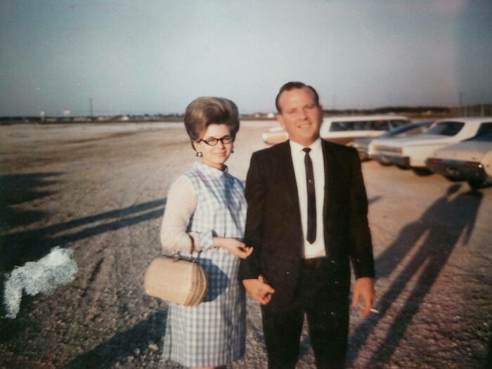 1960s fashion look with a woman in a checkered dress and beehive hairstyle, man in a suit, classic cars in the background.