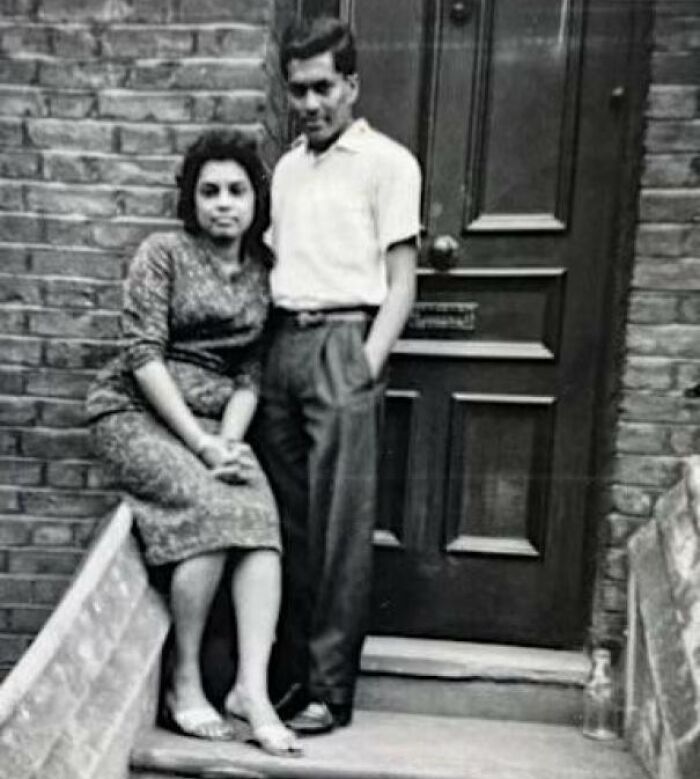 A couple in 60s fashion, with the woman in a patterned dress and the man in a white shirt, standing outside a brick building.