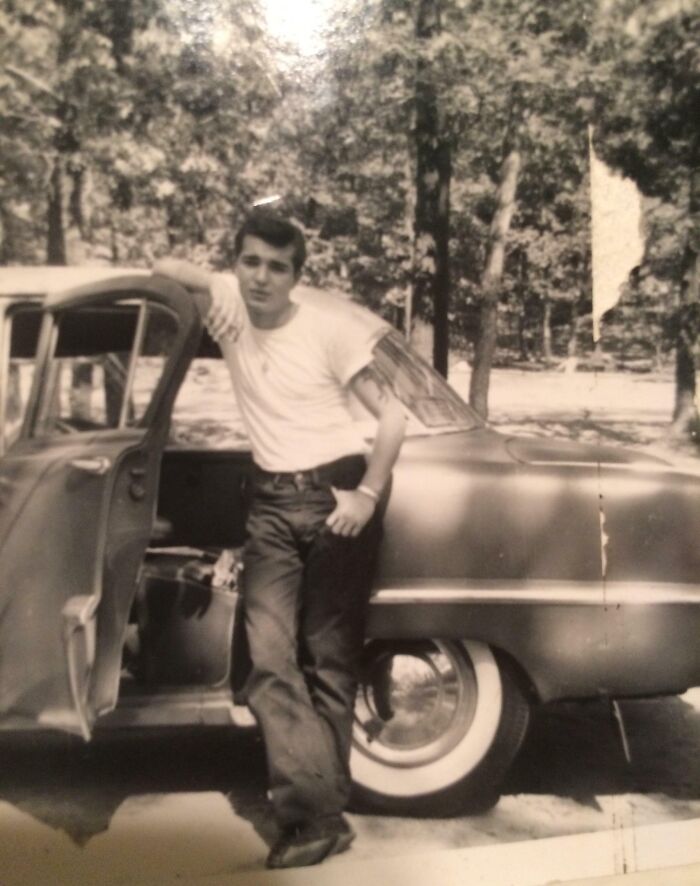 Vintage 60s fashion: man in white tee and jeans leaning on a classic car outdoors.